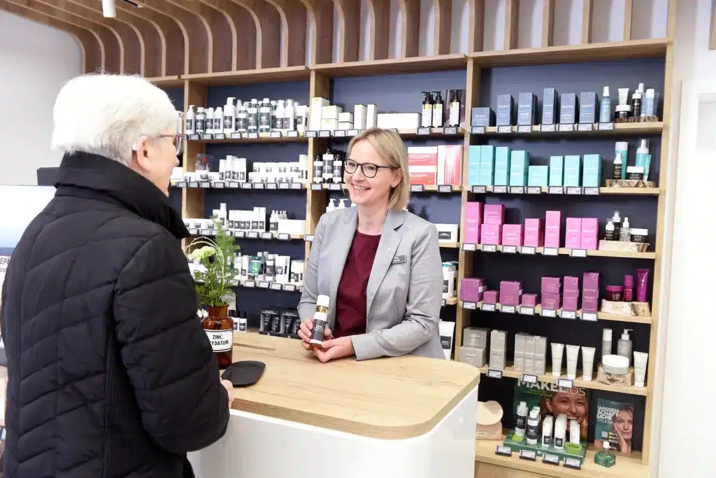 Ein lächelnder Mitarbeiter steht hinter der Theke, hält ein Hautpflegeprodukt in der Hand und unterhält sich mit einem Kunden in einer schwarzen Jacke. In den Regalen hinter ihnen stehen verschiedene Schönheits- und Hautpflegeprodukte.