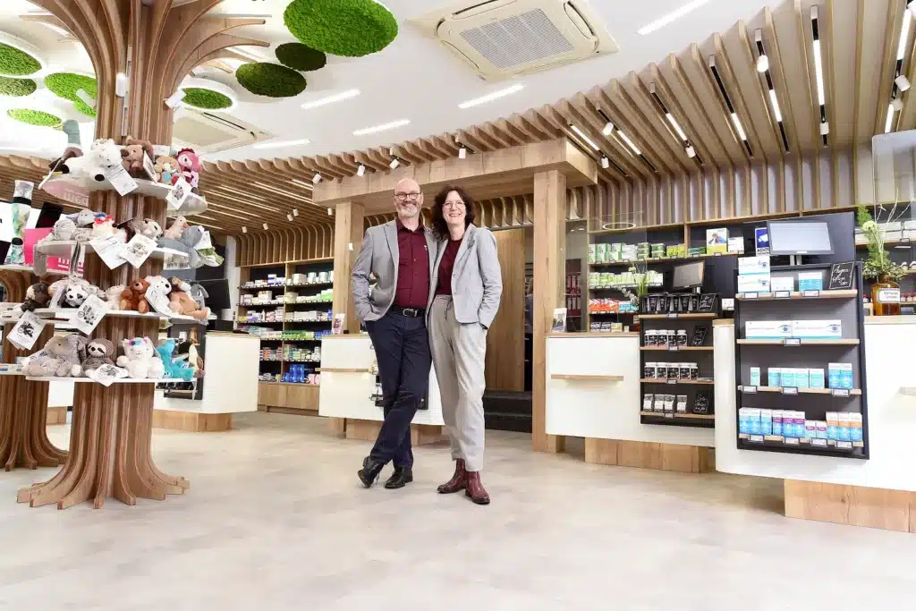 Ein Mann und eine Frau lächeln und posieren gemeinsam in einer modernen Apotheke, umgeben von Regalen mit Produkten und einem Ausstellungsbaum mit Stofftieren. Die Einrichtung besticht durch Holzelemente und runde grüne Deckenplatten.