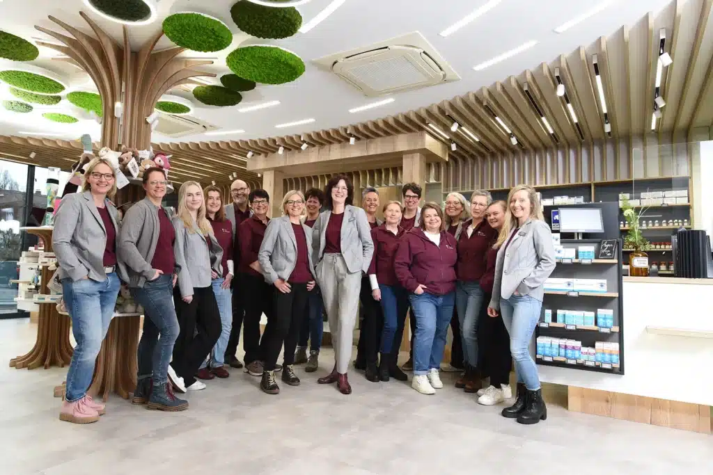 Eine Gruppe von dreizehn lächelnden Menschen, die kastanienbraune Oberteile und graue Blazer oder Freizeitkleidung tragen, steht gemeinsam in einem modernen, hell beleuchteten Geschäft mit Dekorelementen aus Holz und in Grün. Im Hintergrund sind Regale mit Produkten zu sehen.