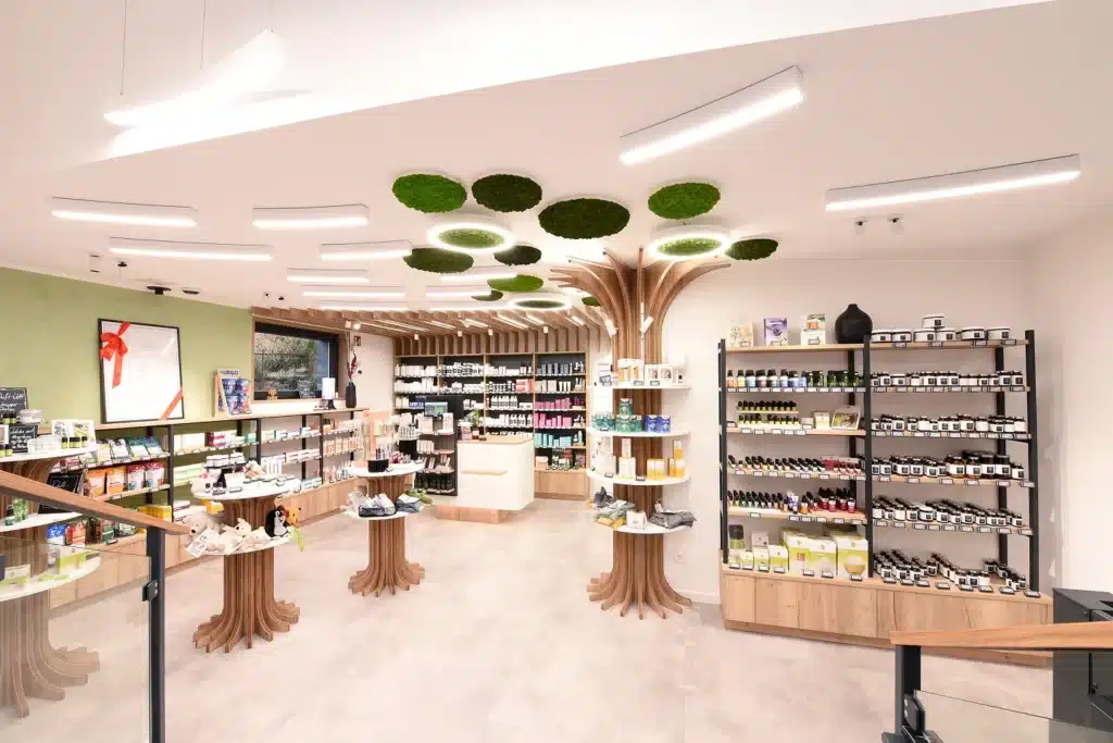 Moderne Apotheke oder Wellness-Geschäft mit heller Beleuchtung, Ausstellungsregalen aus Naturholz in Form von Baumstämmen, grünen Moosdekorationen an der Decke und Regalen, die mit Gesundheitsprodukten, Nahrungsergänzungsmitteln und Hautpflegeartikeln bestückt sind.