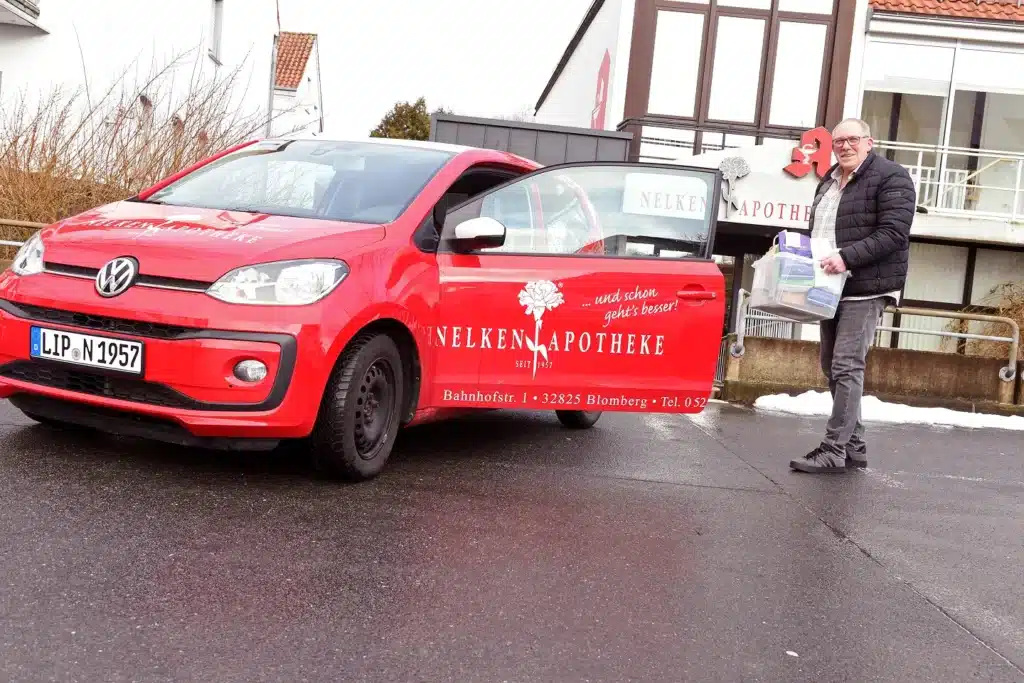 Ein Mann steht neben einem roten Volkswagen, auf dessen Seite „Nelken Apotheke“ steht, und hält eine Schachtel und eine Tüte in der Hand. Das Auto steht auf einer nassen Straße vor einem Apothekengebäude.
