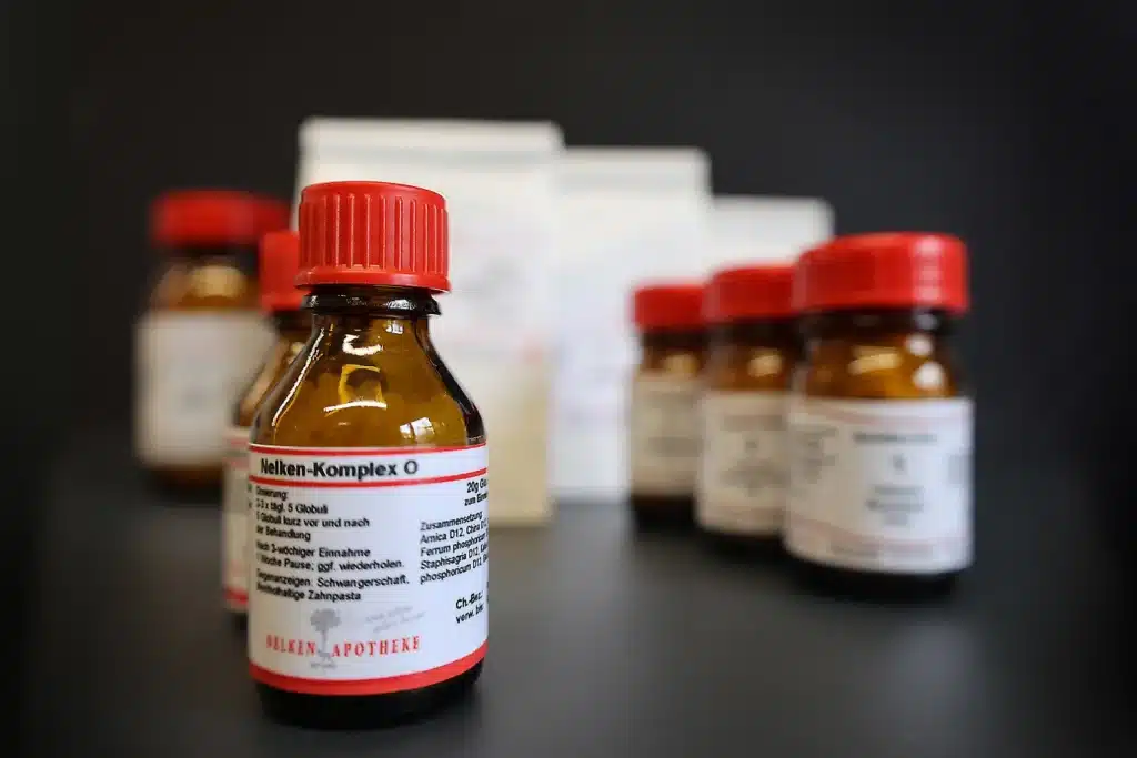 Auf einer schwarzen Fläche sind kleine braune Glasflaschen mit roten Verschlüssen und weißen Etiketten angeordnet. Auf dem Etikett der vorderen Flasche steht „Nelken-Komplex“, daneben ist ein unauffälliger Hinweis „Vorbestellung“ zu sehen. Im Hintergrund sind weitere Flaschen und weiße Päckchen verschwommen zu erkennen.