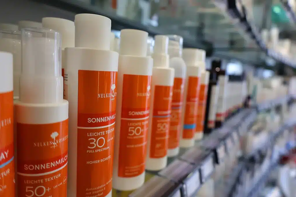 In einem Ladenregal stehen Flaschen mit Sonnencreme mit orangefarbenen Etiketten in Reih und Glied, auf denen verschiedene Lichtschutzfaktoren und Produktdetails angegeben sind; einige sind mit speziellen Etiketten für die Vorbestellung versehen.