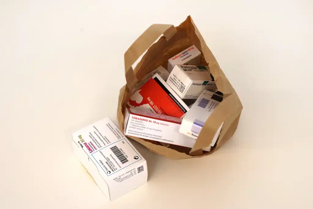 Auf einer weißen Fläche liegt eine braune Papiertüte, gefüllt mit verschiedenen Schachteln mit verschreibungspflichtigen Medikamenten; eine Schachtel steht außerhalb der Tüte, bereit für die Vorbestellung.